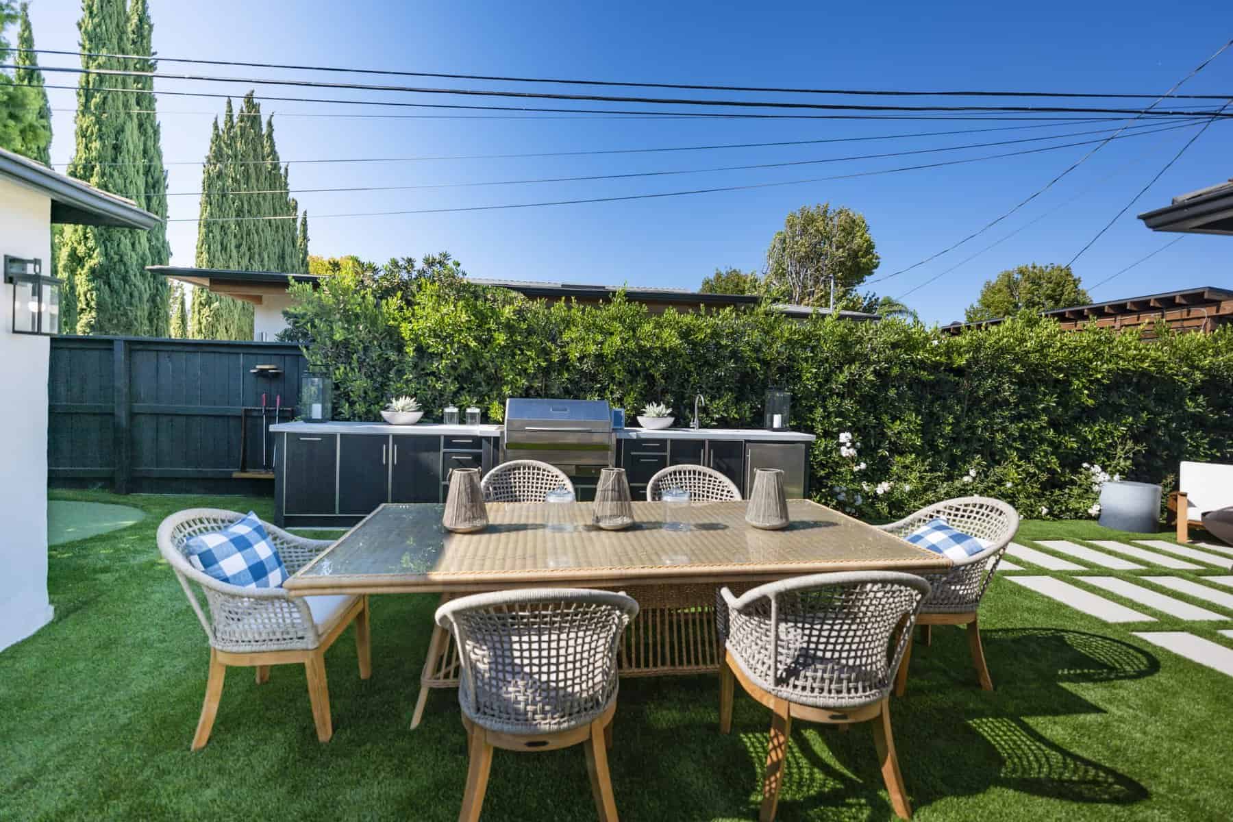 Modern backyard with outdoor kitchen, dining table, wicker chairs, and lush greenery