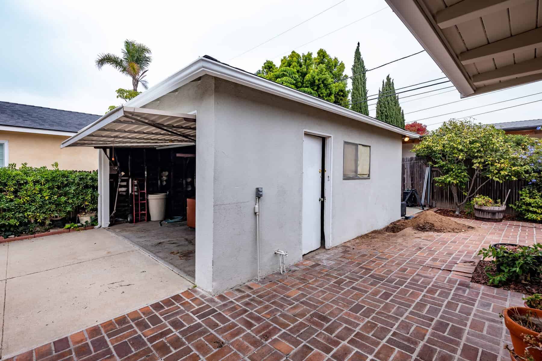 Detached garage with an open door, white exterior, and brick-paved backyard surrounded by greenery