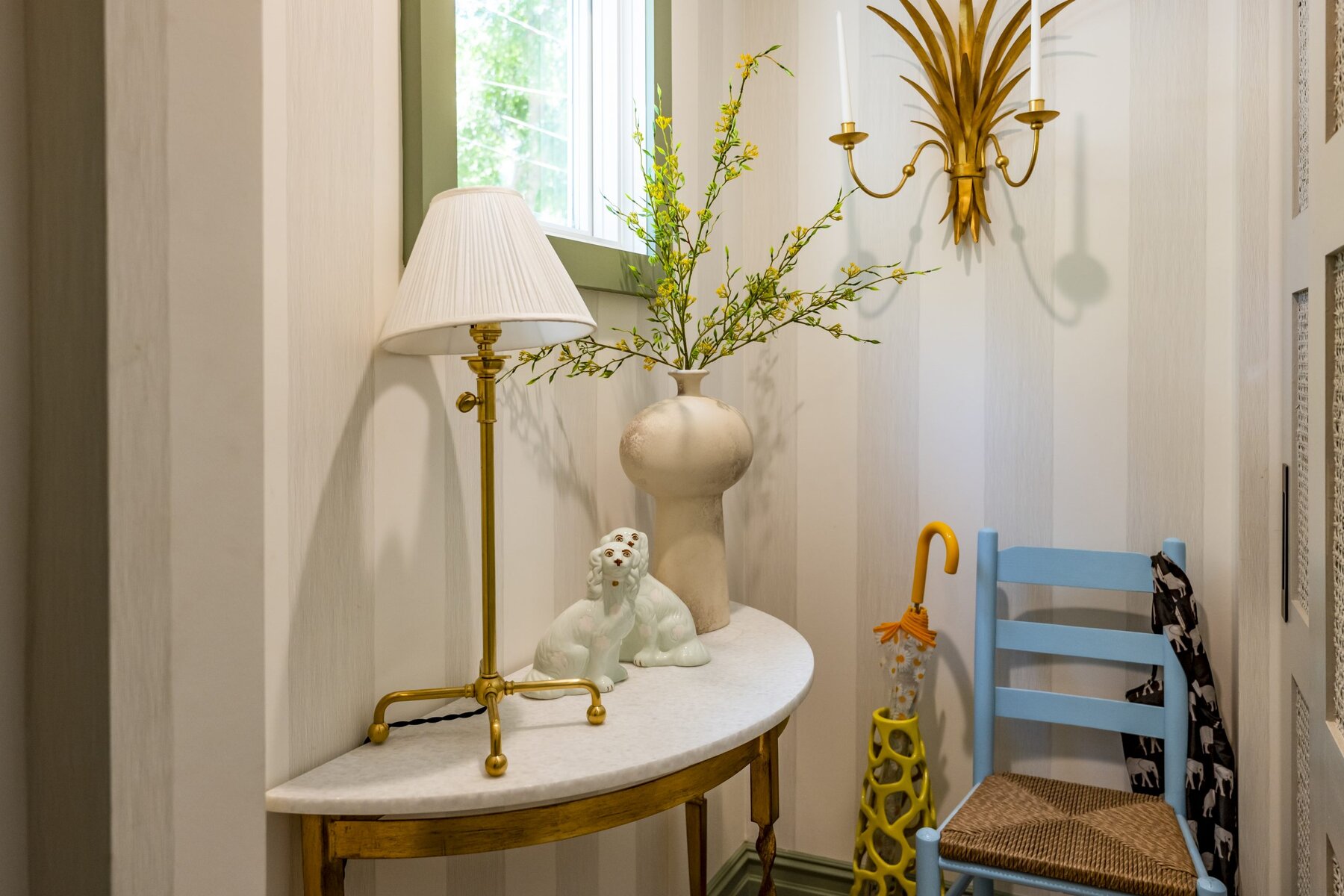 Entryway with a marble table, golden lamp, floral arrangement, blue chair, umbrella, and wall sconce