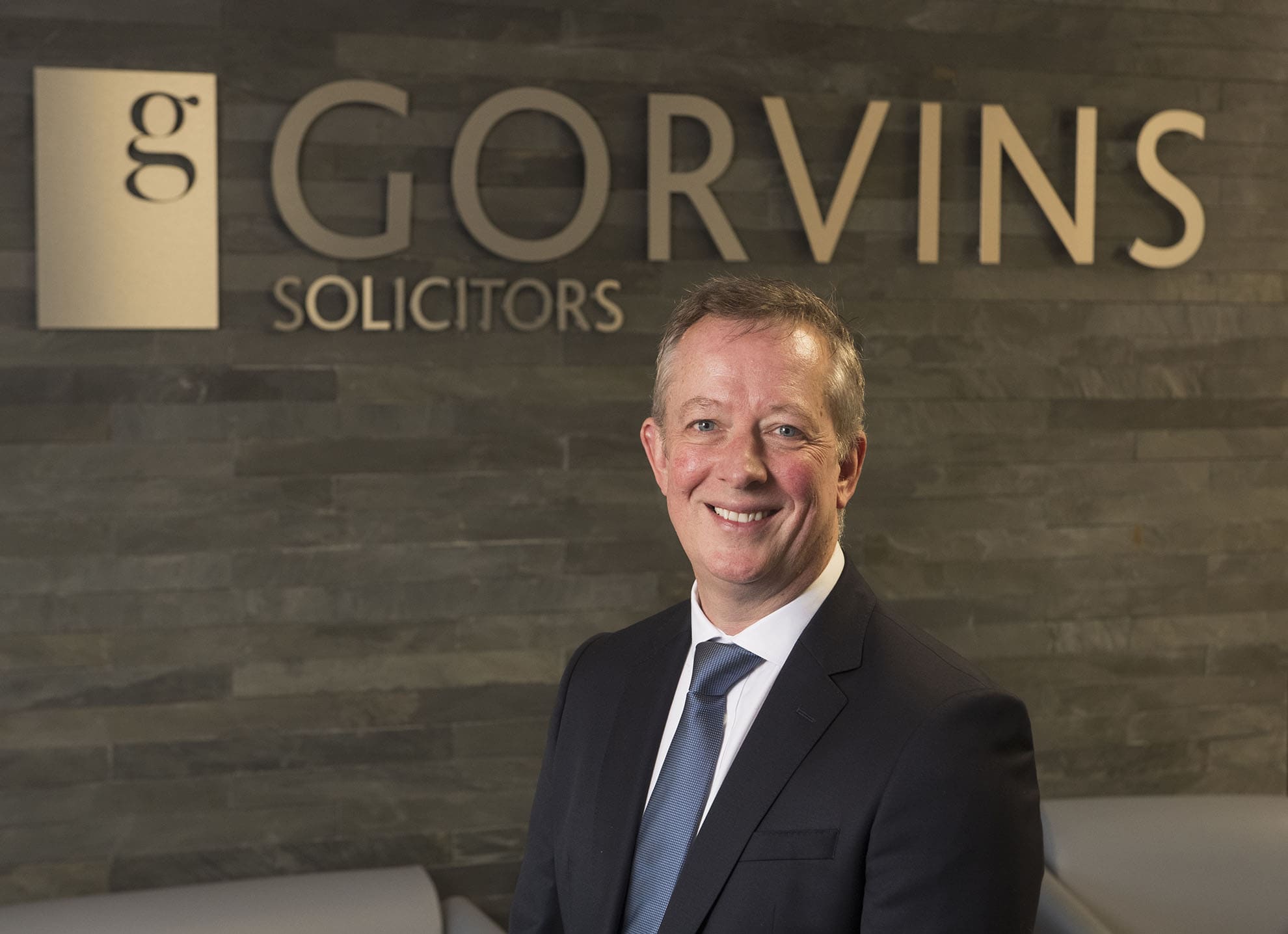 Experienced solicitor in a suit smiling at the Gorvins Solicitors office with modern tile wall background.