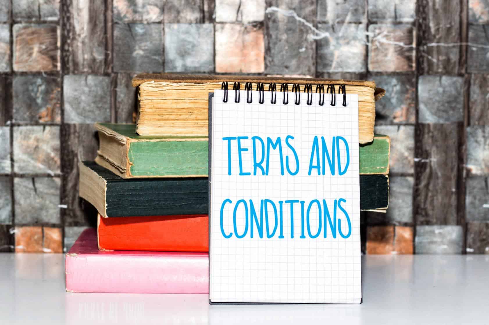 Terms and conditions sign on a notepad placed in front of a stack of old books against a rustic brick wall.
