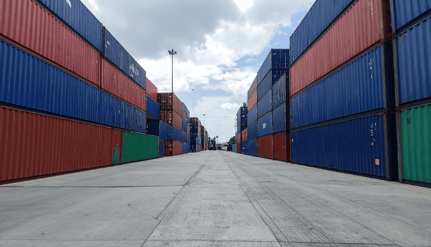 Stacked shipping containers at a busy port or storage yard, with a concrete ground and partly cloudy sky.