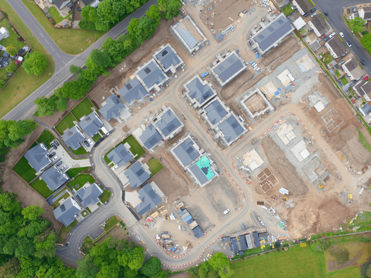 Aerial view of new residential construction site with several houses in various stages of building, surrounded by greenery and streets.