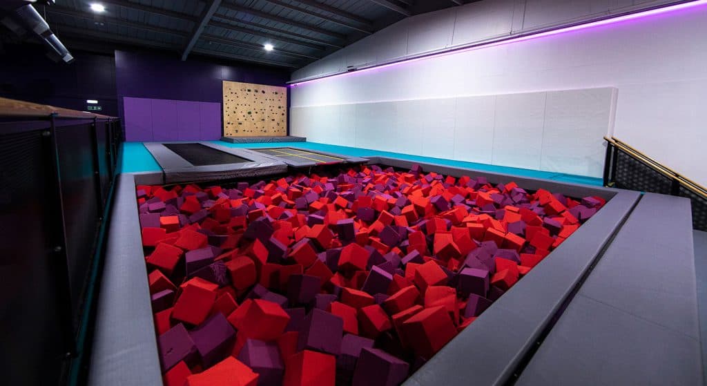 Soft play area with red and purple foam cubes under a trampoline and climbing wall in an indoor recreational facility.