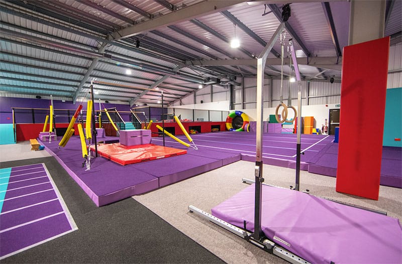 Colourful indoor fitness and gymnastics training facility with soft mats, climbing frames, rings, and obstacle courses for children and adults.