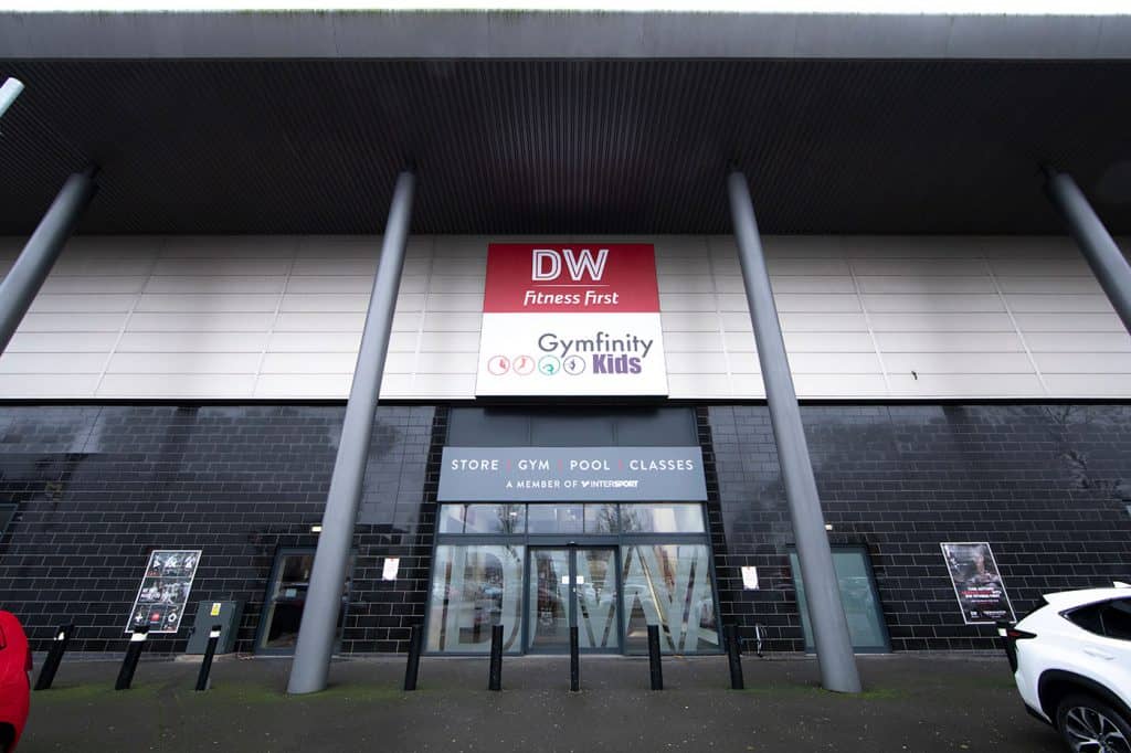 Fitness centre entrance with signage for DW Fitness First and Gymfinity Kids, modern building façade, front view with black brick and glass doors, parking area in front.