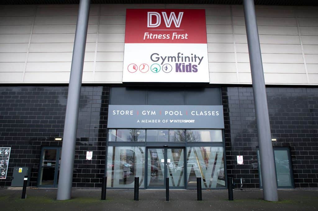 Gym entrance signage for DW Fitness First and Gymfinity Kids with store, gym, pool, and classes information in black and white on a dark building facade, with large metallic support poles.