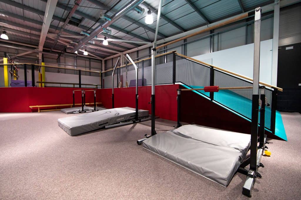 Aerial yoga and fitness studio with soft mats, trapeze bars, and climbing frames in a spacious industrial-style gym with high ceilings and bright lighting.