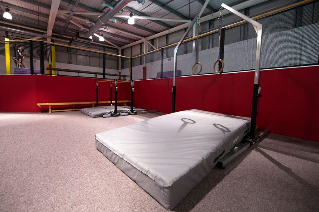 Gymnastics training area with mats and equipment for aerial exercises and strength training in a sports facility, bright lighting and industrial ceiling.