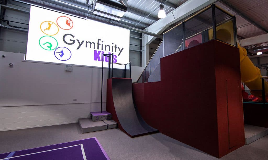 Gymfinity Kids indoor play area with a large digital screen displaying the logo, featuring a slide and climbing structures suitable for children.