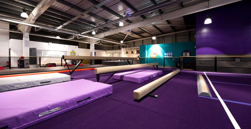 Flexible gymnastics training area with mats and vaulting equipment in a spacious, well-lit indoor sports facility with purple flooring and high ceilings.