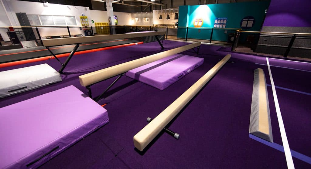 Gymnastics training area with padded mats, balance beams, and spotting equipment in a modern indoor sports facility.