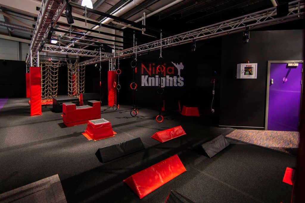A ninja-themed obstacle course with red and black padding, climbing ropes, gymnastic rings, and various challenge zones for physical training and entertainment.