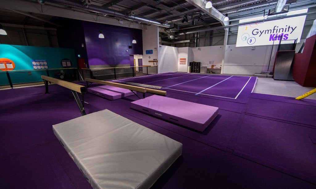 A gymnastics training area with purple mats, a balance beam, uneven bars, and foam pads in a modern indoor gym for kids and beginners.