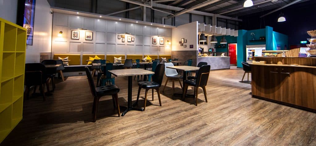 Brightly lit modern cafe interior with a mix of colourful chairs, wooden tables, wall art, and a counter area for ordering food and drinks, creating an inviting atmosphere.