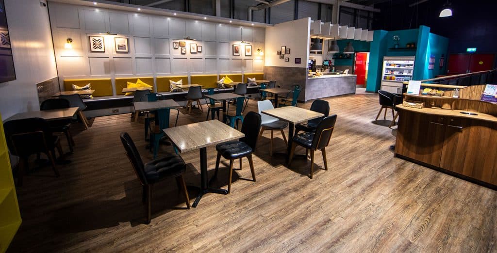 Cozy modern café interior with wooden flooring, colourful chairs, wall art, and a counter area with a coffee machine and supplies, creating a welcoming environment.