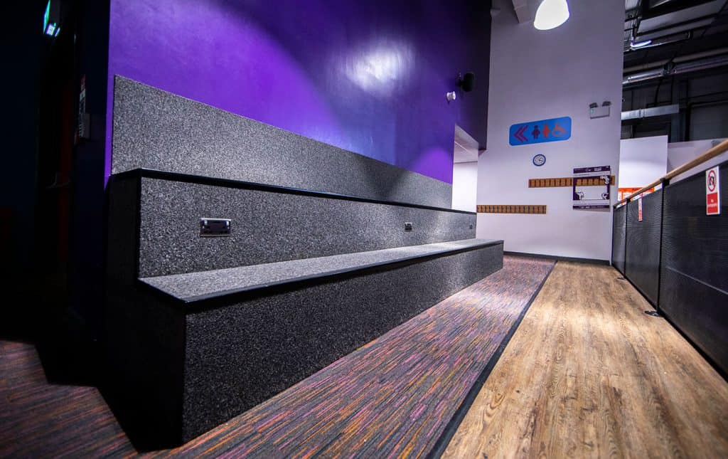 A modern indoor space with a purple accent wall, wooden flooring, and a granite seating area. Signage for restrooms and accessibility is visible, suggesting it could be a public or recreational facility.