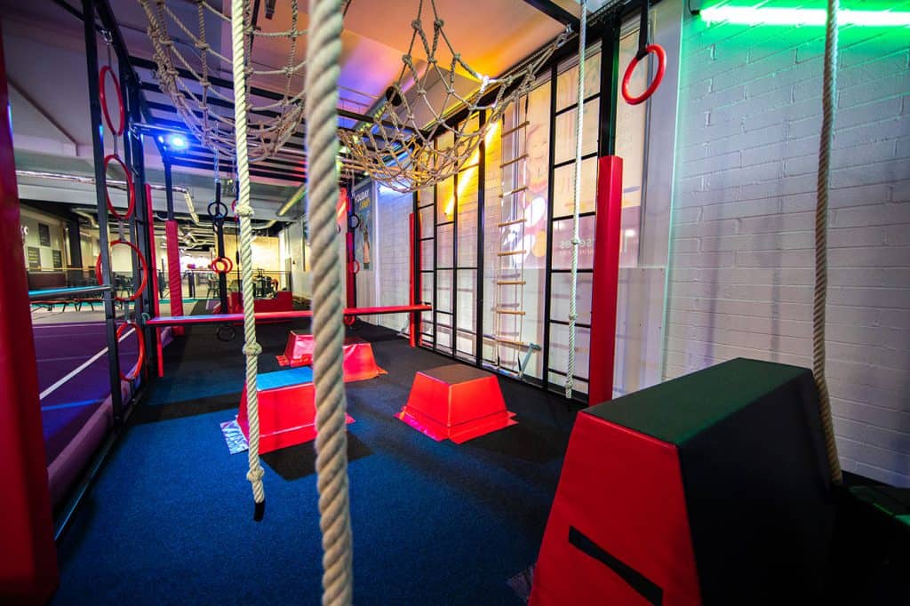 Colourful indoor children's play area with climbing ropes, padded obstacles, and rings in a modern entertainment centre. Bright lighting and vibrant equipment create an engaging environment for kids.