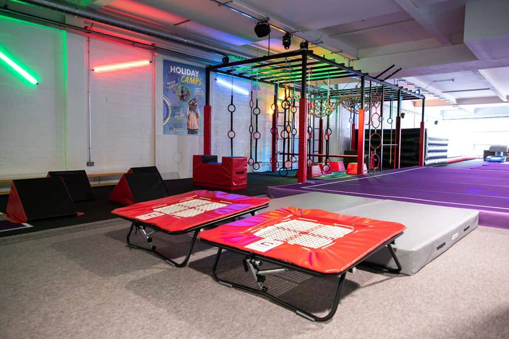 AstroTurf trampolines in an indoor kids' activity centre with colourful climbing frames and neon lighting for active play and fitness fun.