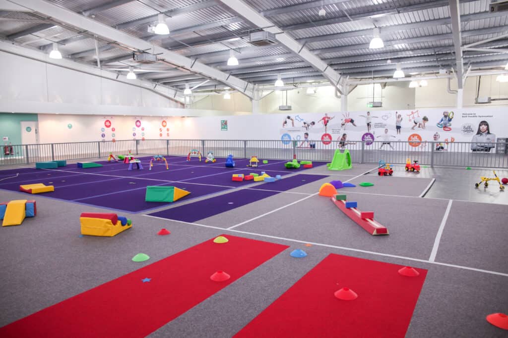 Bright indoor children's play area with colourful soft play equipment, slides, and toys inside a spacious gymnasium designed for kids' activities and gymnastic practice.