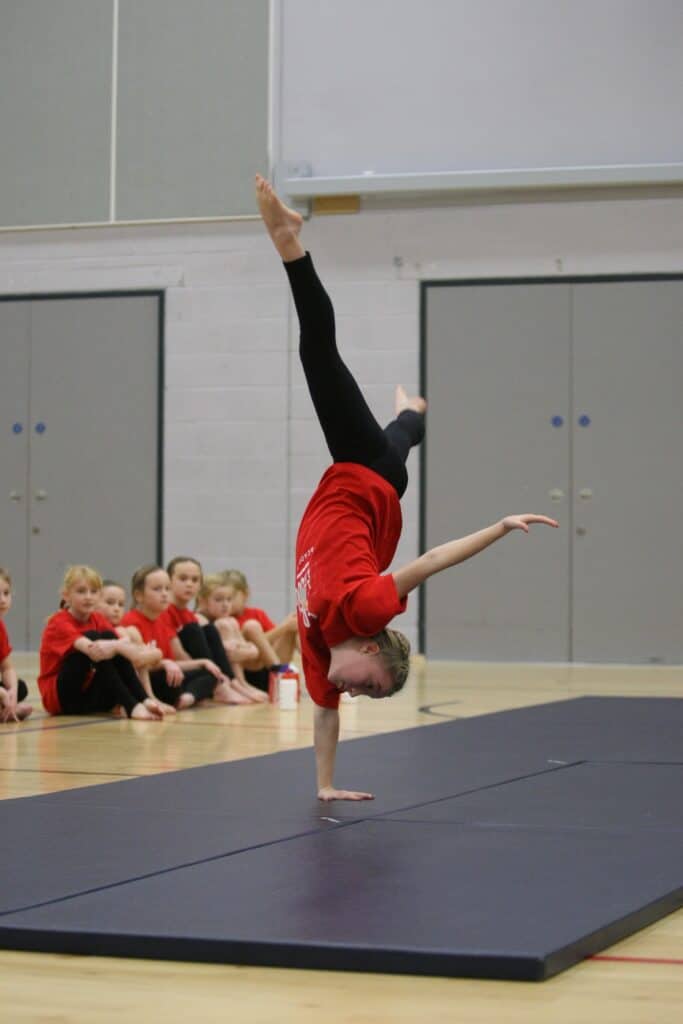 Handstand gymnast performing a one-handed handstand on a black mat in a gymnasium with an audience of young gymnasts in red uniforms watching.