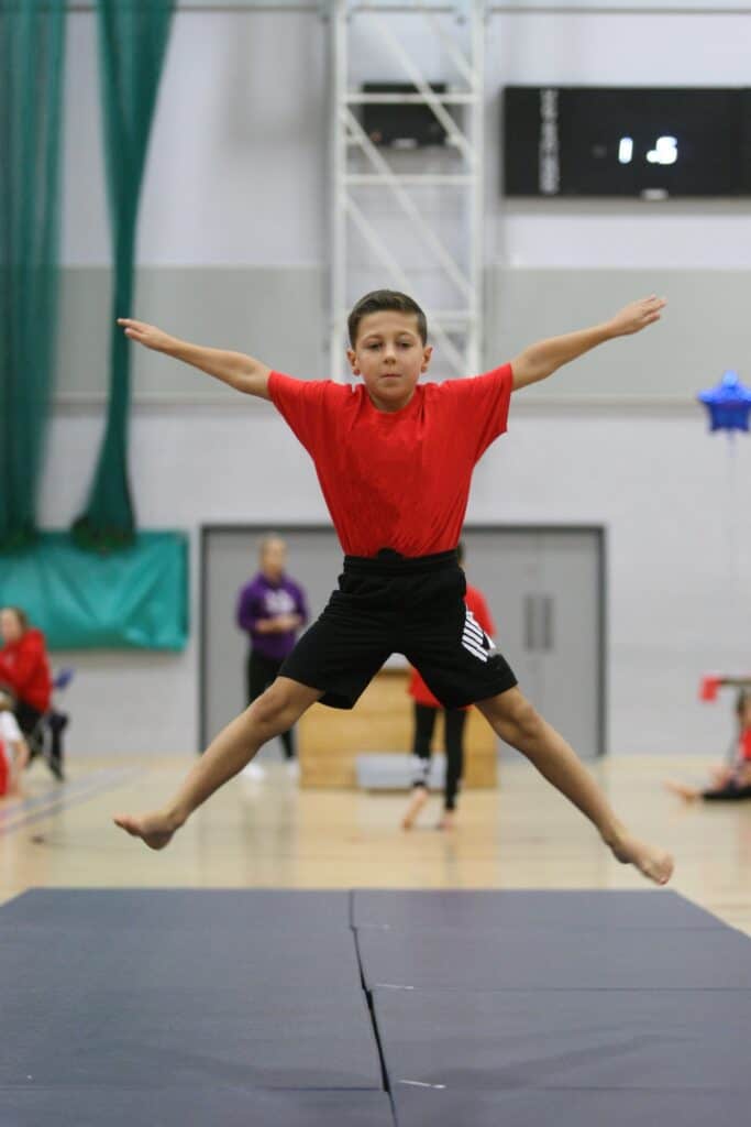 Boy performing gymnastics jump on black mat in indoor sports hall.