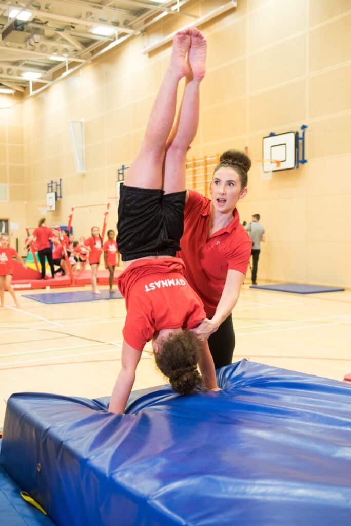 Dynamic gymnastic coaching session for young athletes in a sports hall. The instructor is assisting a girl with a handstand, demonstrating flexibility and strength training for gymnastics. Multiple children in matching red shirts are practicing with various equipment in the background.