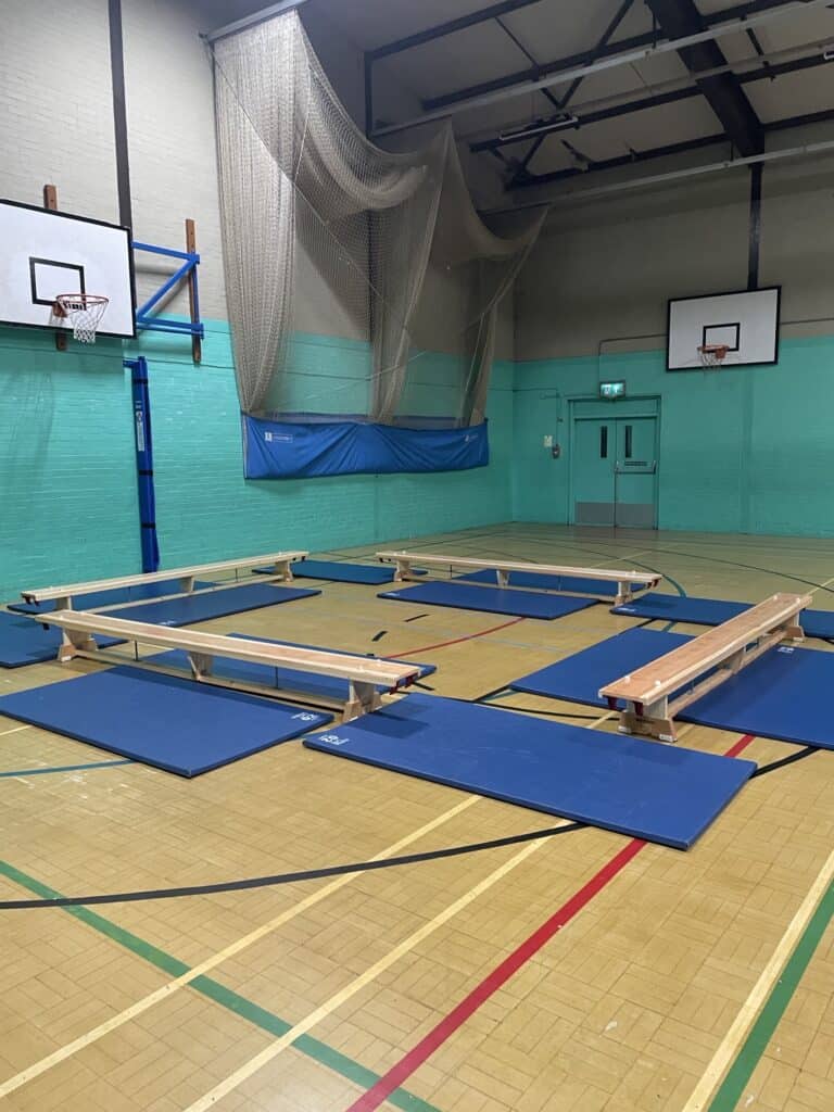 Climbing training area with padded mats, benches, and nets in an indoor sports hall. Suitable for gymnastics or physical education activities. Brightly coloured walls and basketball hoops visible in the background.