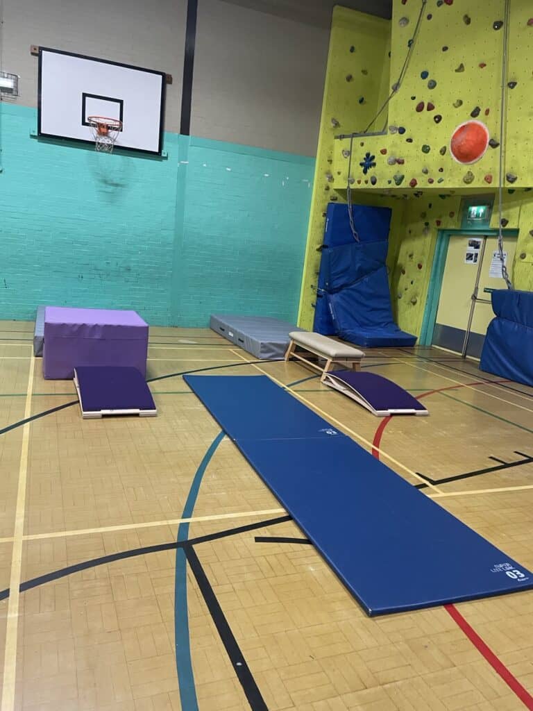 Climbing gym with padded mats, a rock wall, basketball hoop and coloured markings on a wooden sports hall floor, ideal for indoor fitness and recreation activities.