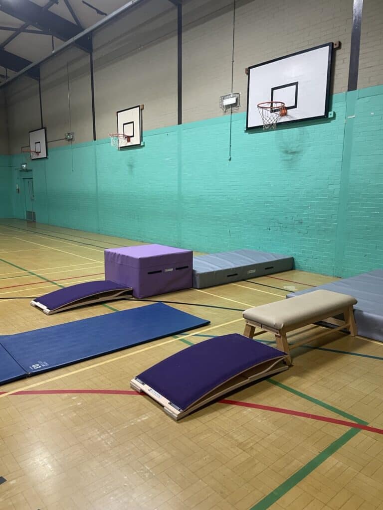 Gymnastics training area with mats and vaulting equipment in a school sports hall. Multiple basketball hoops on the vibrant teal painted brick wall add to the space's multi-sport functionality.