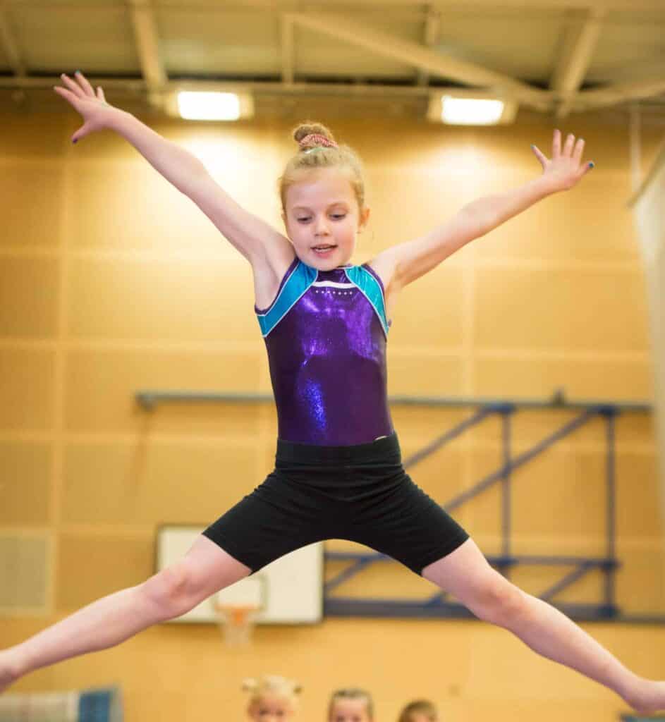 Dynamic young gymnast in a purple and blue leotard performing a star jump in an indoor gymnasium.