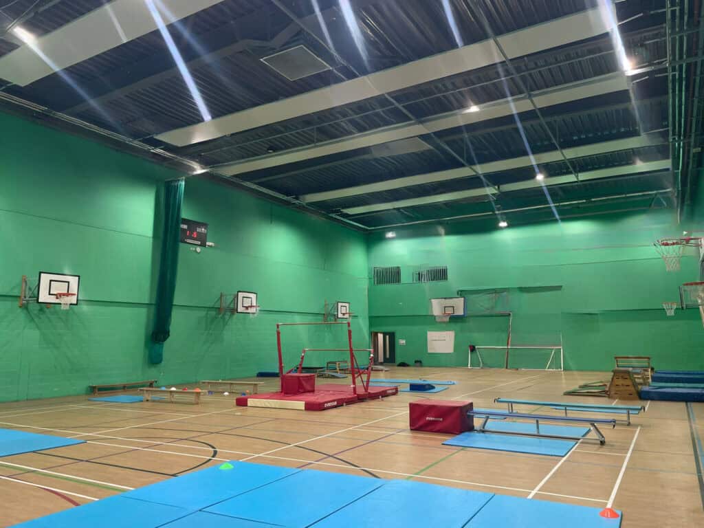 Brightly lit indoor sports hall with green walls, equipped for gymnastics and basketball, featuring mats, vaults, hoops, and training equipment for athletic activities.