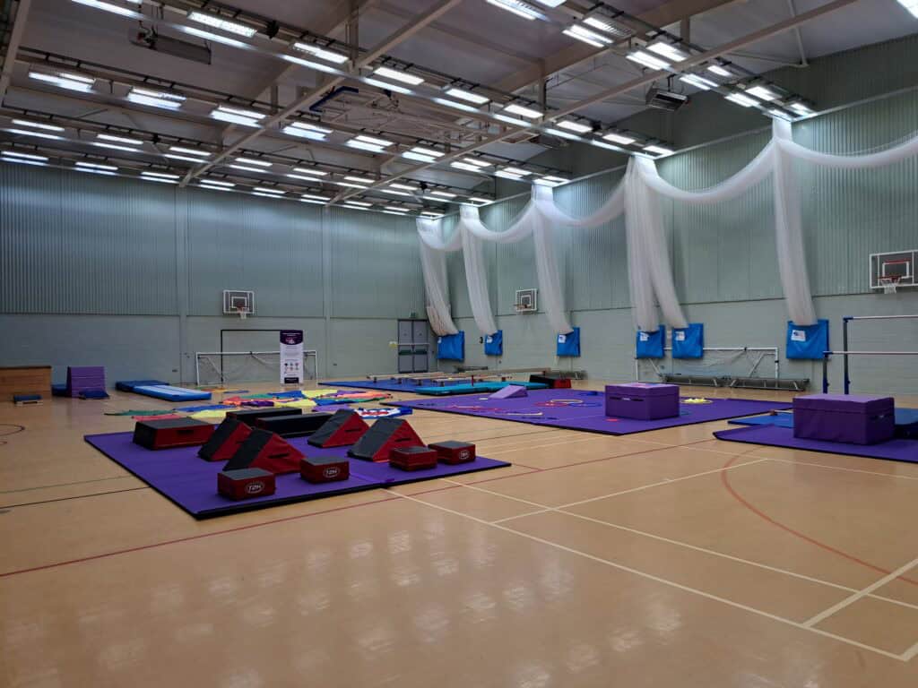 Gymnastics training facility with mats, equipment, and basketball hoops in a spacious indoor sports hall, suitable for gymnastics and physical education activities.