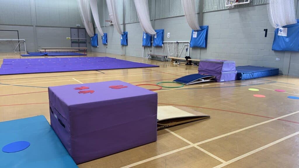 Softplay gymnasium equipped with purple mats, foam blocks, hoops, and colourful circular stepping stones, set up for children's activity and gym classes in an indoor sports hall.
