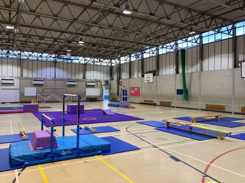 Gymnastics training facility with padded mats, balance beams, and various gym equipment inside a spacious indoor sports hall with high ceilings.