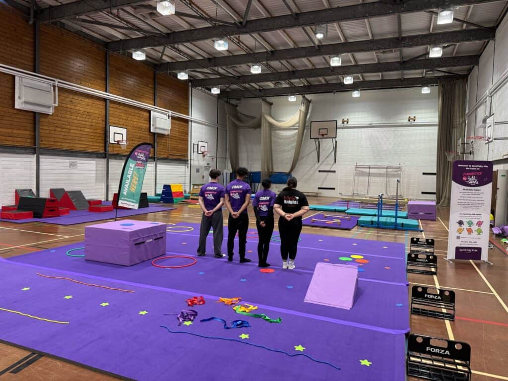 Brightly lit indoor gymnasium set up for children's gymnastics or acrobatic activities, with purple mats, soft play equipment, hoops, and coaches briefing participants.