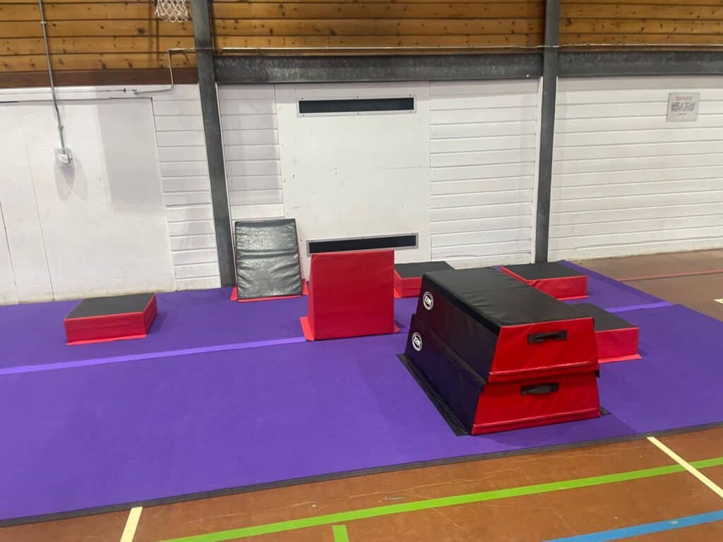 Padded obstacle course set up on a purple gym mat inside a sports hall or gymnasium. The setup includes various padded blocks and platforms for parkour or training exercises.