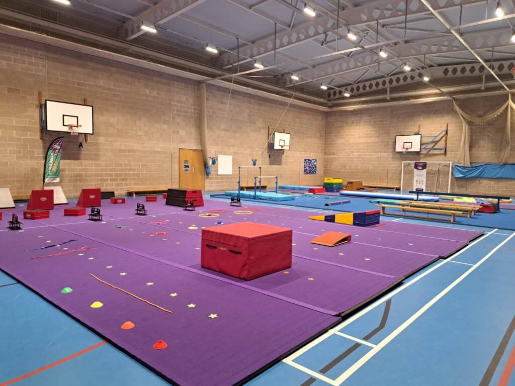 A spacious indoor gymnasium set up for gymnastics training, featuring colourful mats, balance beams, rings, and basketball hoops on the walls, with ample space for diverse physical activities.