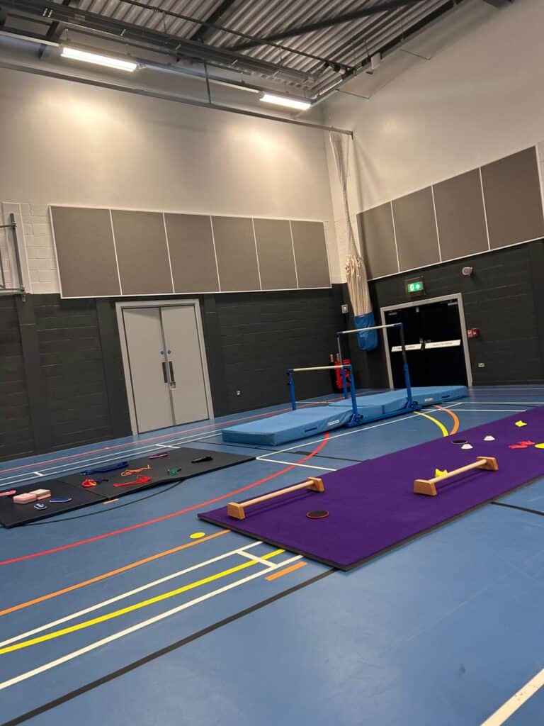 Gymnastics training area with mats, uneven bars, and various equipment in an indoor sports hall with high ceilings and bright lighting.