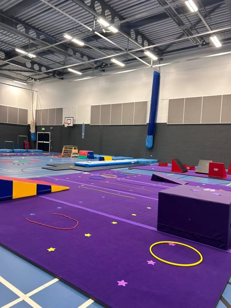 Bright indoor gymnastics training area with colourful mats, balance beams, hoops, and climbing equipment in a spacious gymnasium.