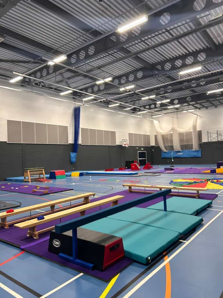 Bright indoor gymnasium with colourful gymnastics mats, benches, and equipment for training and physical activities, illuminated by overhead lighting in a spacious, organized environment.