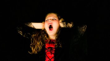 A young girl with curly hair covering her ears, screaming or shouting with a distressed expression against a black background.