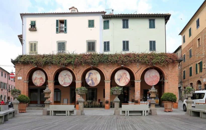 Fari artistici sulle arcate di un edificio storico a partire dalla terrazza, con affreschi di figure femminili, nel centro di una piazza di città italiana.