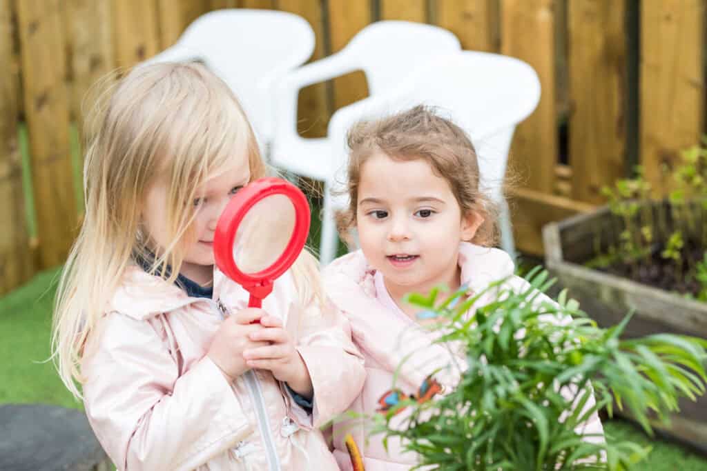 Little girls exploring outdoors at Thrive Childcare, engaging in playful learning activities with a magnifying glass, in a safe, nurturing environment perfect for early childhood development.