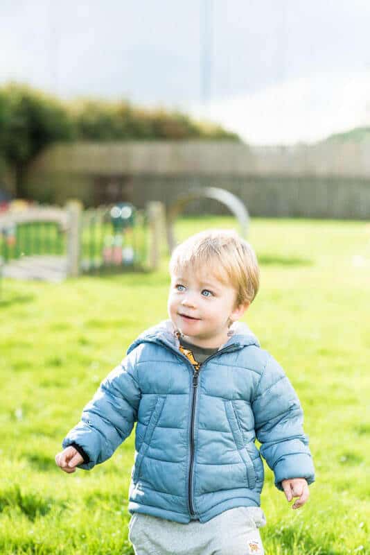1. Young child playing outside in a secure childcare playground, wearing a blue jacket and enjoying outdoor activities at Thrive Childcare.