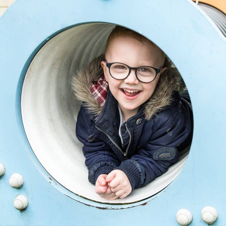 Bright smiling child with glasses playing inside a playground tube, promoting early childhood development and outdoor play activities at Thrive Childcare.