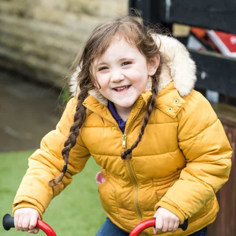 Bright smiling young girl with pigtails riding a red tricycle outdoors, demonstrating joyful childhood experiences at Thrive Childcare.