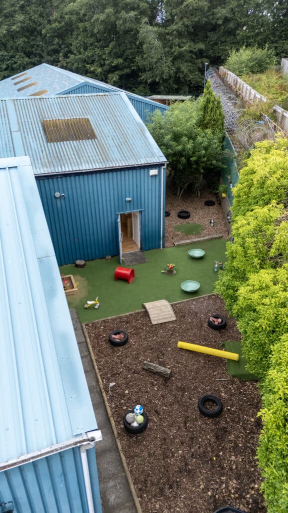 Bright blue outdoor play area at Thrive Childcare with artificial grass, toys, tyres, and wooden bridge, surrounded by lush greenery for outdoor activities and exploration.
