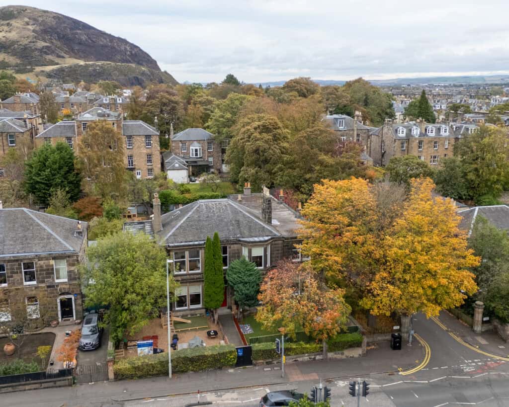 Bright aerial view of a charming childcare centre surrounded by vibrant autumn trees, nestled in a residential area with traditional stone buildings and scenic hills in the background, emphasising a nurturing environment.