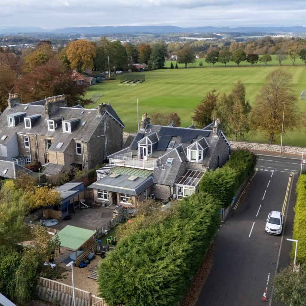 Bright aerial view of Thrive Childcare's modern building set against a spacious green playing field, surrounded by trees and quiet neighbourhood streets, emphasising a safe, outdoor play environment for children.